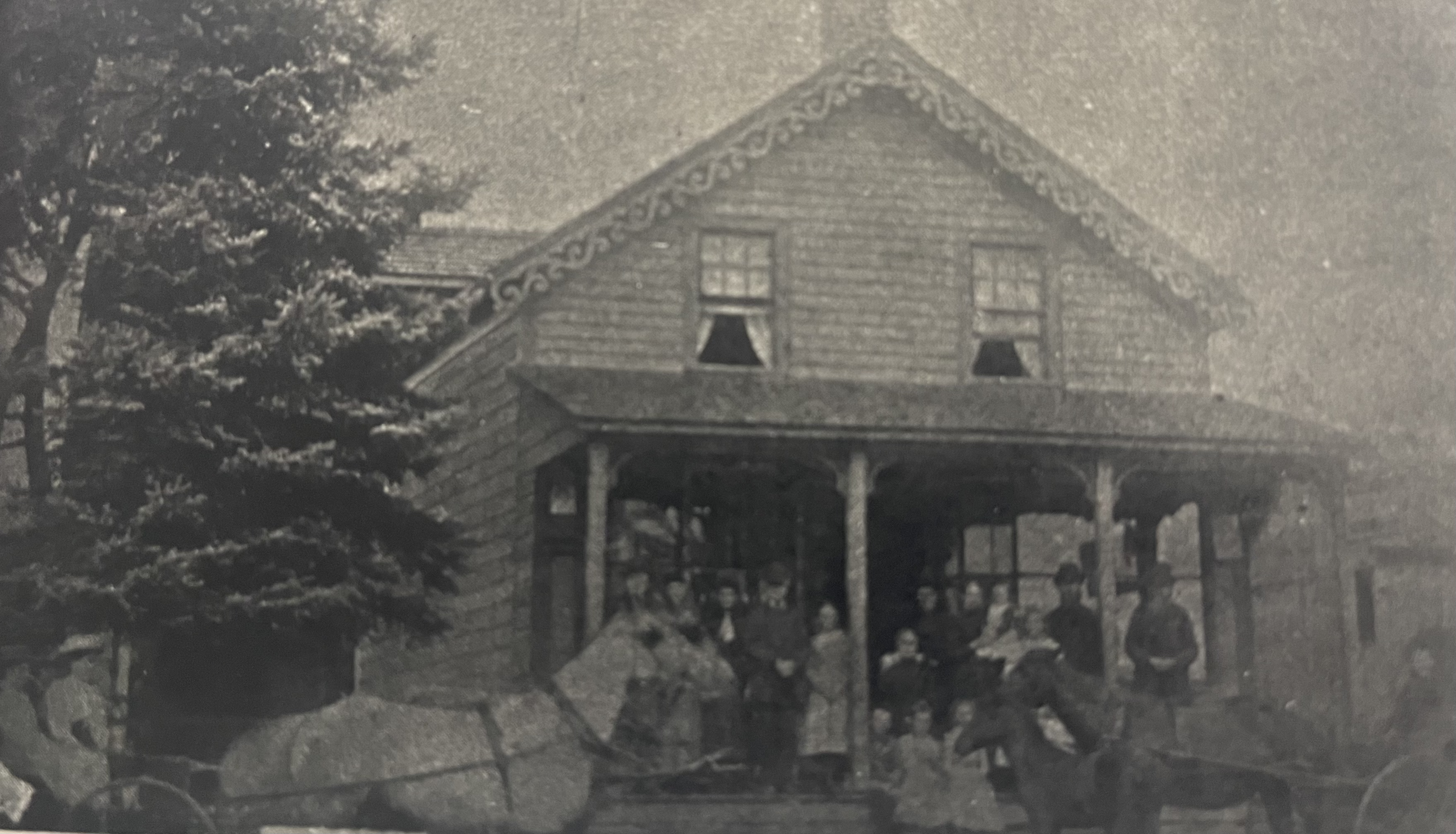 An aged photograph of a community gathering on the porch of an old farm house.